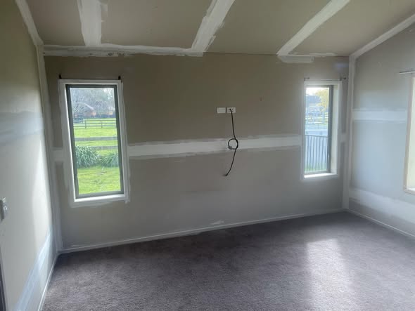 Kitchen / living room mid-build with raw gib walls and paper-tape joins, doorway through to a finished kitchen behind